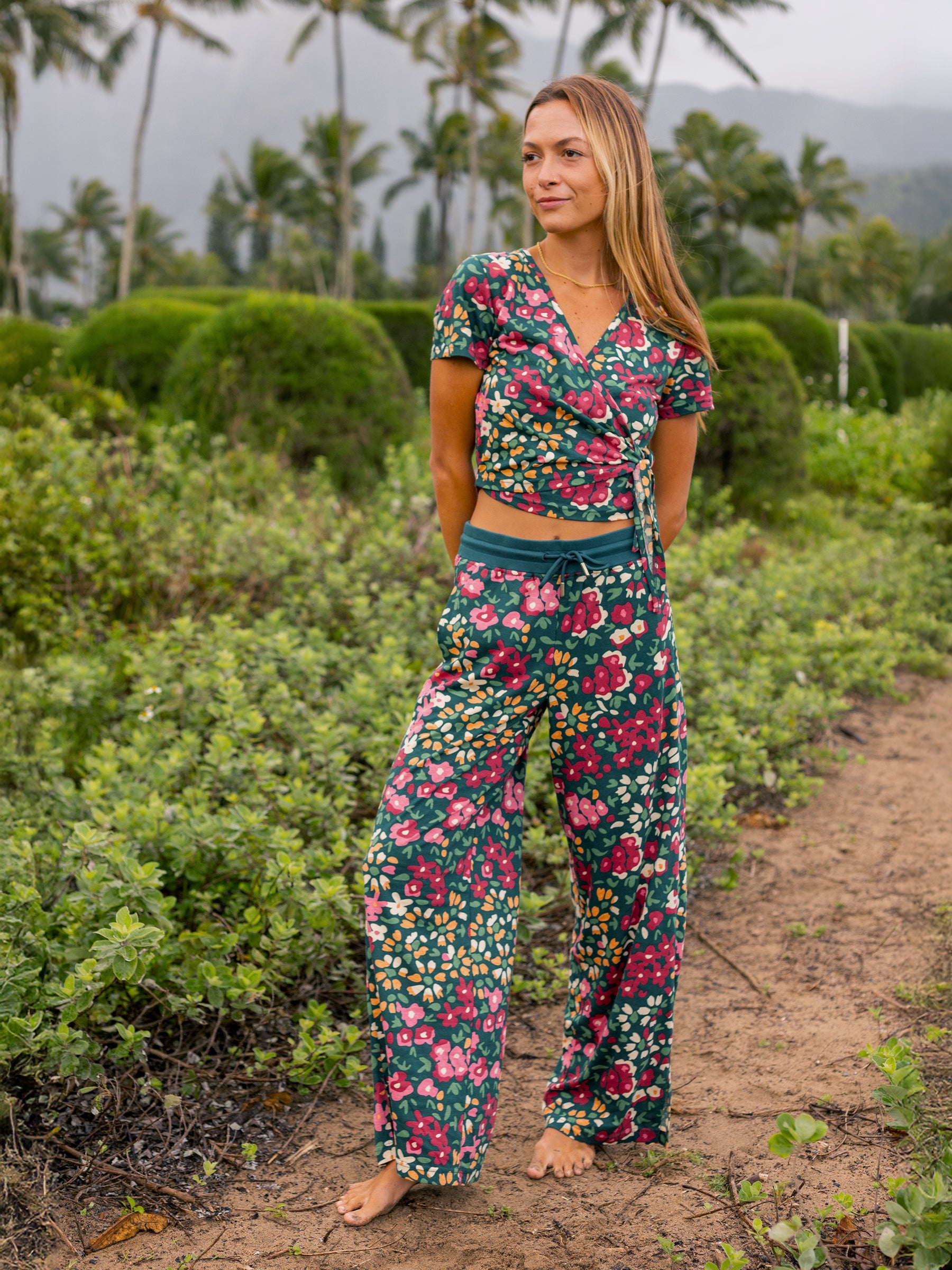 Woman in a green organic cotton wide-leg pants and matching floral top, standing on a dirt path.